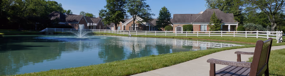 A peaceful outdoor scene at The Villas at St. Therese - Assisted Living featuring a pond with a water fountain, a white fence surrounding the area, several buildings in the background, green grass, trees, and a wooden bench on a paved walkway.