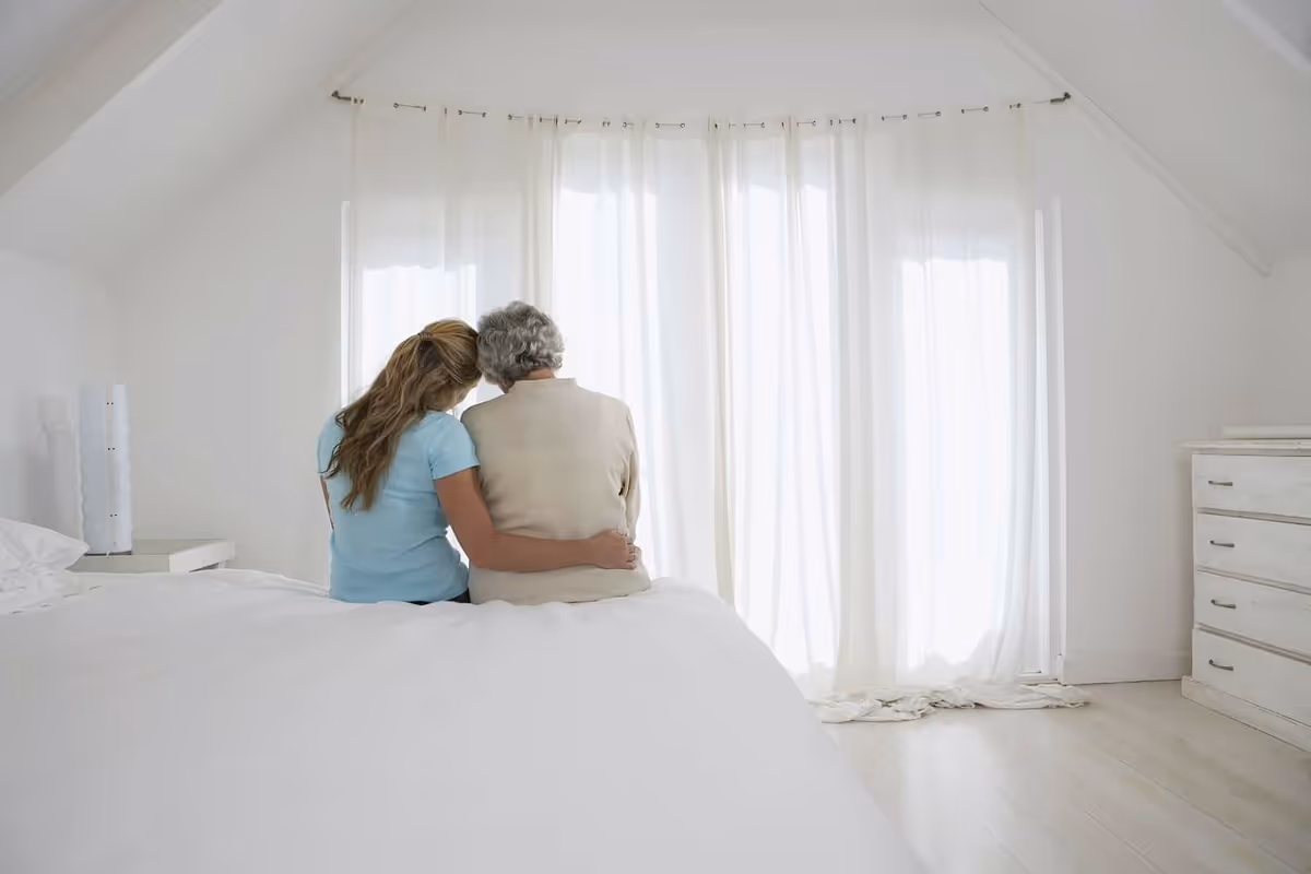 A woman with long hair in a blue shirt sits on a bed with her arm around an elderly person with gray hair, both facing away from the camera towards sheer white curtains covering a window in a bright, white bedroom.