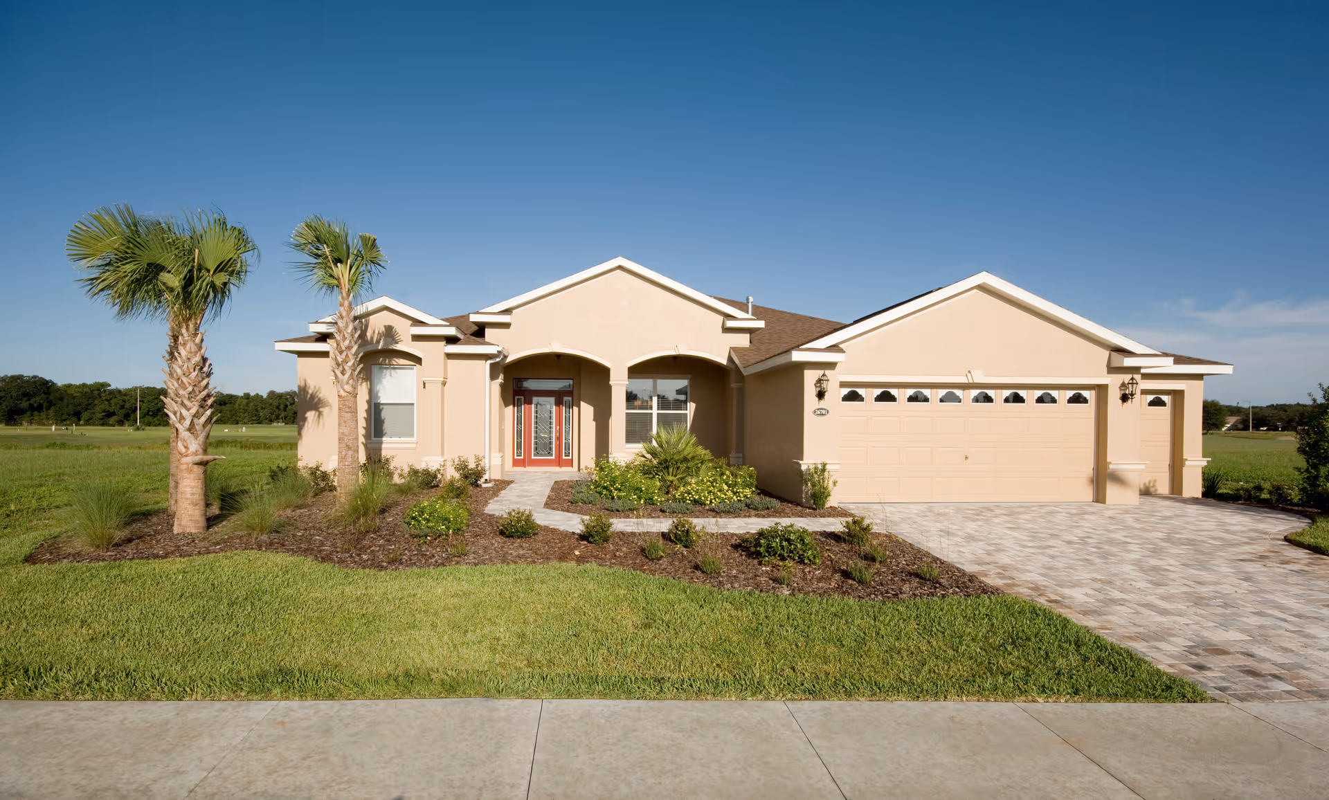 Single-story beige house with a two-car garage, palm trees, and a landscaped front yard under a clear blue sky.