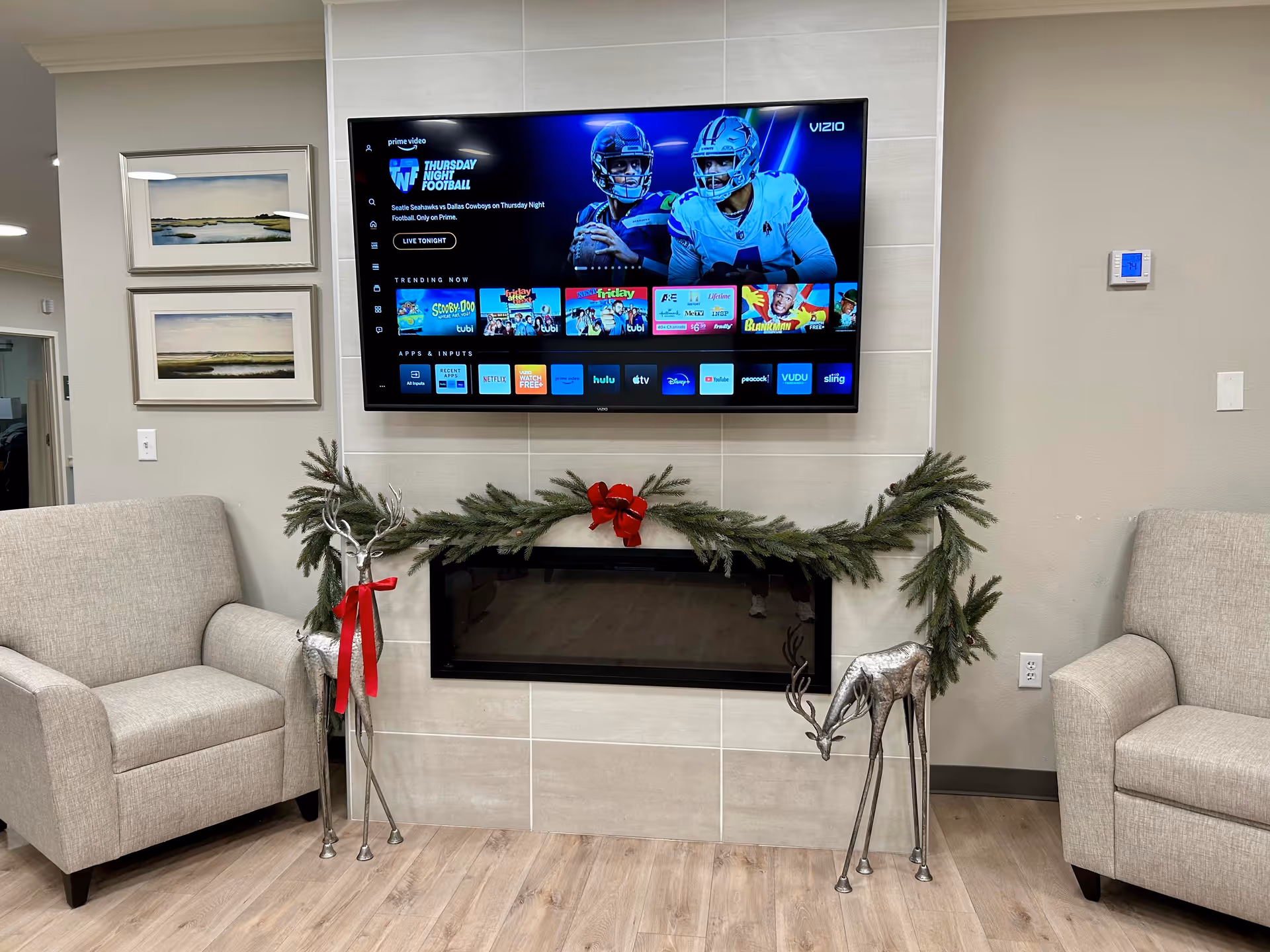 A cozy living room area with two beige armchairs on either side of a modern electric fireplace decorated with a green garland and red bow. Above the fireplace is a wall-mounted flat-screen TV displaying a streaming service interface with a football game featured. Two silver decorative reindeer with red ribbons stand on either side of the fireplace. Two landscape paintings hang on the wall to the left.
