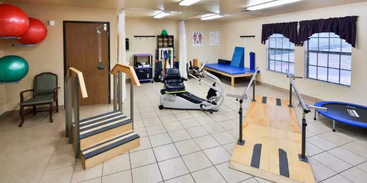 A physical therapy room with exercise equipment including a small staircase with handrails, a ramp with handrails, a recumbent exercise bike, a blue padded therapy table, a small trampoline, and large red and green exercise balls mounted on the wall. There are two windows with dark curtains and anatomical charts on the wall.