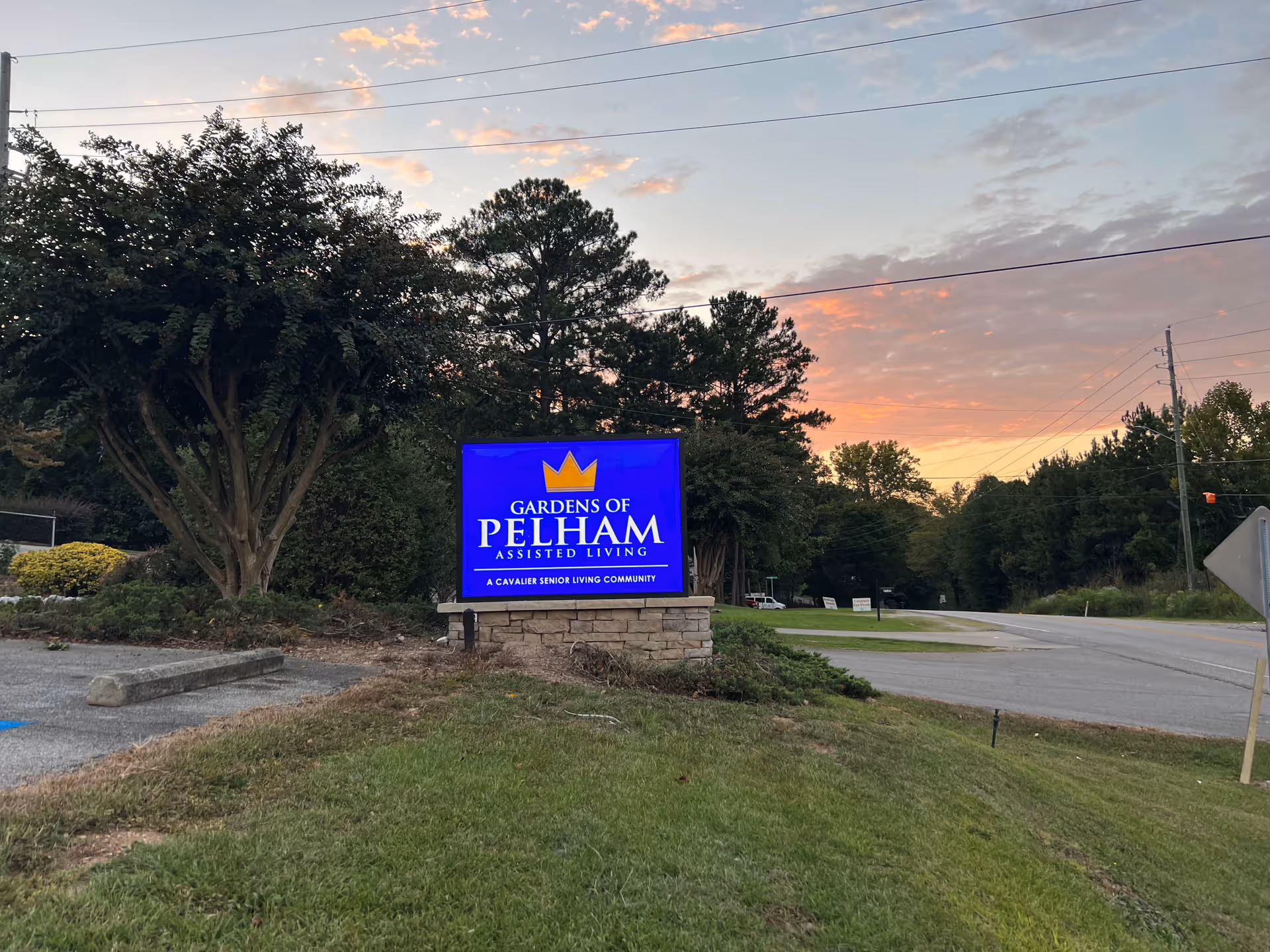 Outdoor view of a blue sign for Gardens of Pelham Assisted Living, situated on a stone base with grass and trees around it, under a sky with scattered clouds at sunset.