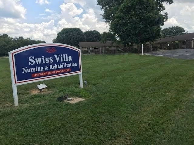 Outdoor view of Swiss Villa Nursing & Rehabilitation facility sign on a grassy lawn with a building and parking lot in the background under a partly cloudy sky.