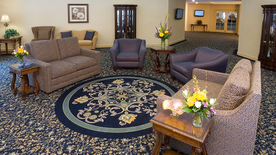 A senior living community lounge area with patterned carpet and a circular floral rug in the center. The room features two patterned sofas, two purple armchairs, wooden side tables with flower arrangements, a beige loveseat, a striped armchair, a glass-front cabinet, and a hallway leading to double doors.