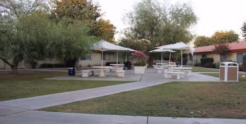 Outdoor courtyard with umbrellas, concrete picnic tables, and a paved walkway by a building.