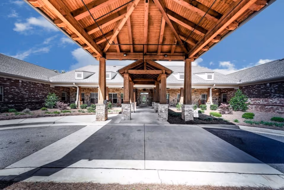 Covered entrance of a brick building with wooden beams and stone pillars, surrounded by landscaped bushes and plants under a blue sky with some clouds.
