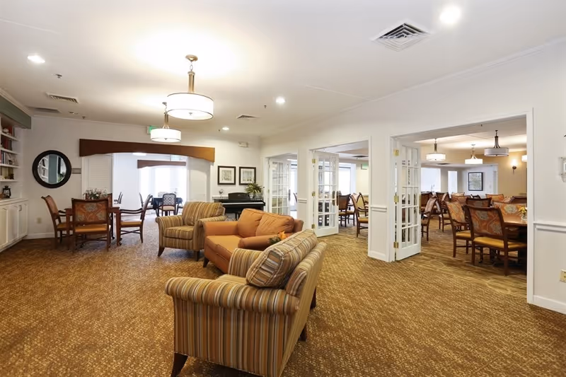 A bright communal lounge with sofas, armchairs and an adjacent dining area in a senior living facility.