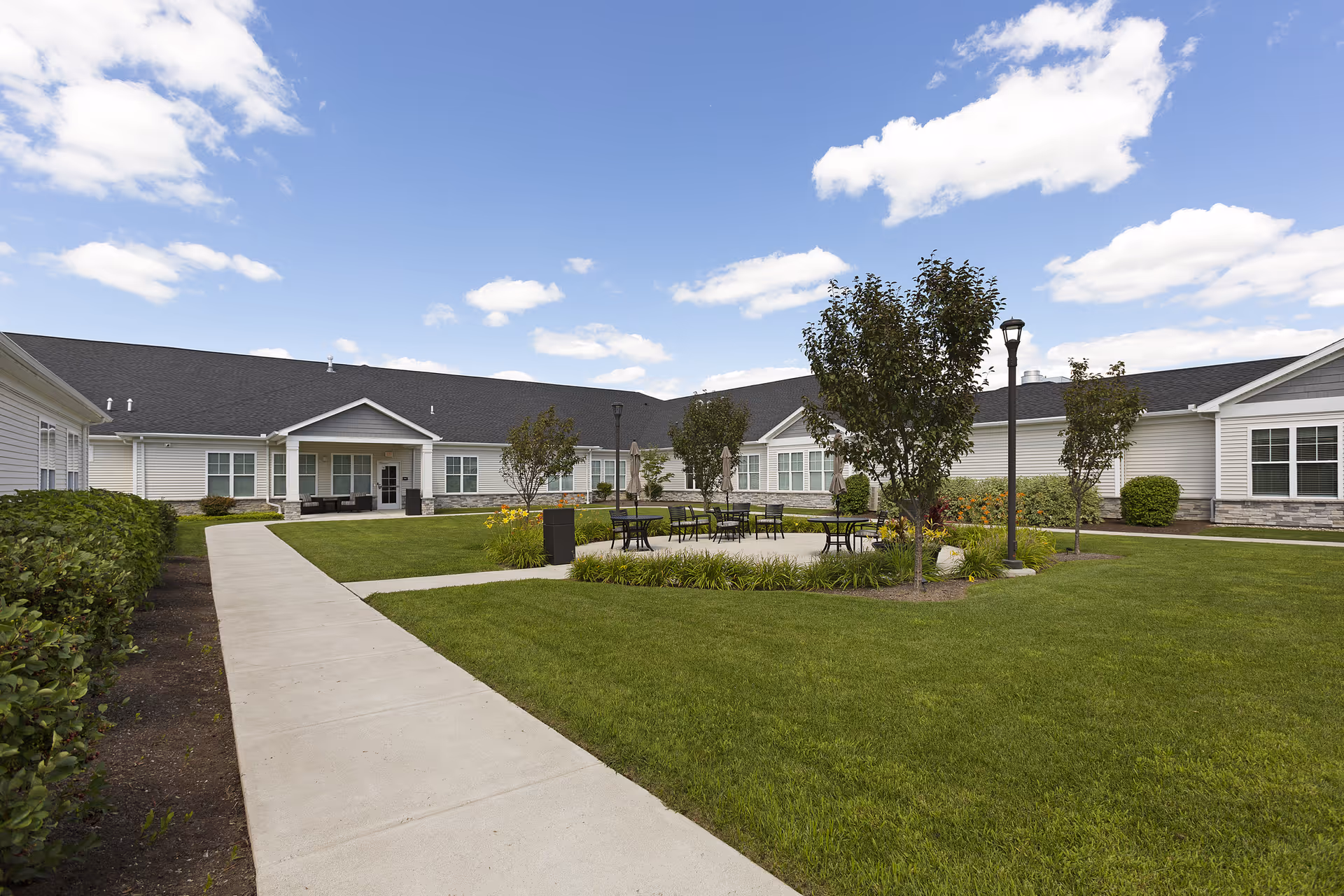 Outdoor courtyard area of a senior living facility with a concrete walkway, green lawn, small trees, and several black metal tables and chairs under a blue sky with scattered clouds. The building surrounding the courtyard is single-story with white siding and a dark roof.