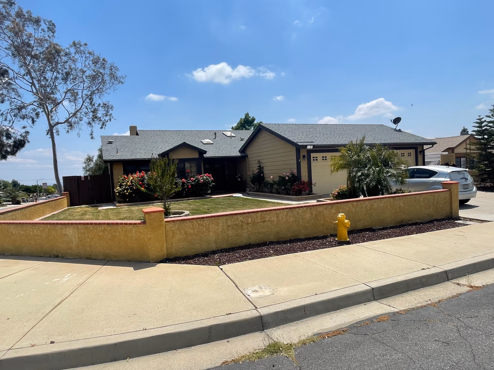 Exterior view of a single-story residential building with a gray roof and beige walls, surrounded by a low yellow stucco wall with red brick trim. The front yard has a small tree, bushes with flowers, and a green lawn. A silver car is parked in the driveway next to the garage. A yellow fire hydrant is visible on the sidewalk in front of the property. The sky is clear with a few clouds.