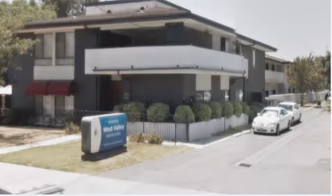 Exterior view of a two-story assisted living facility building with balconies, bushes along the front, a sign that reads 'West Valley Assisted Living', and two white cars parked on the driveway.