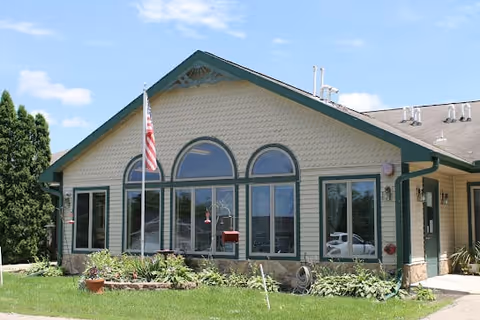 Exterior view of a single-story building with beige siding and green trim, featuring three large arched windows and several rectangular windows. An American flag is mounted on a pole near the building. There are plants and shrubs in front of the building, and a clear blue sky above.