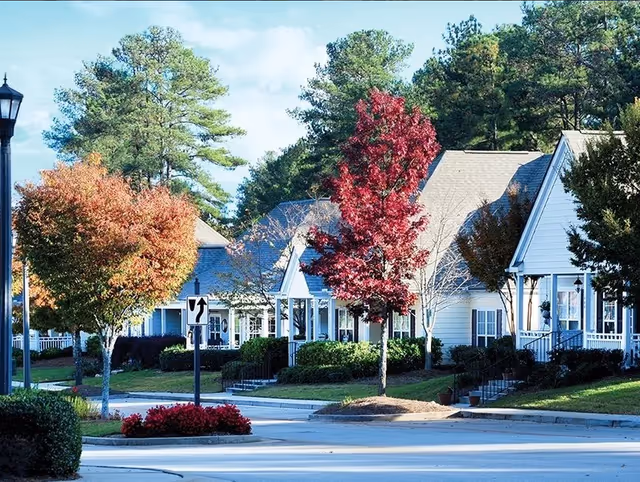 Row of single-story white cottages with porches along a tree-lined street, some trees showing fall colors.