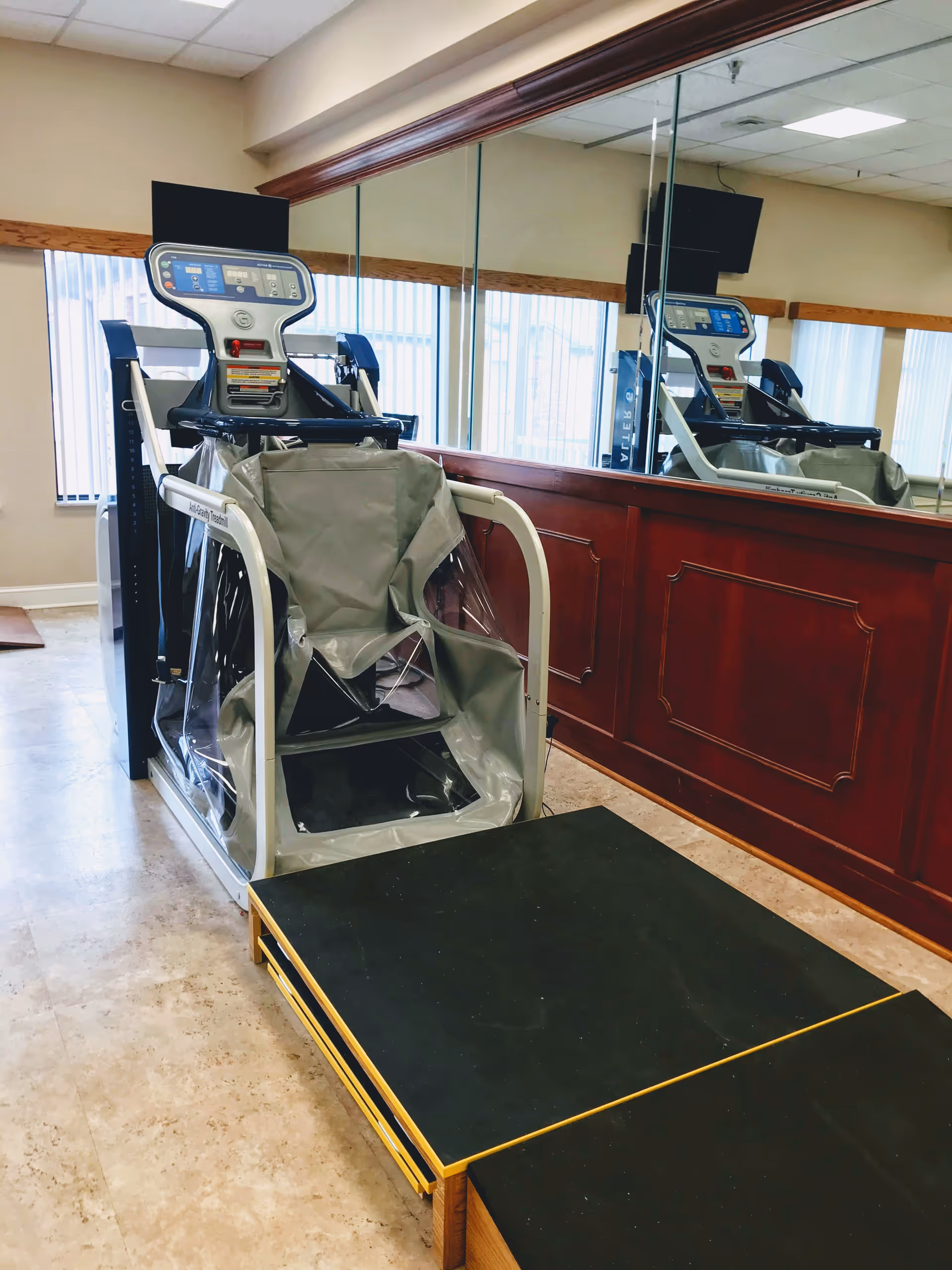 A specialized treadmill with a supportive harness system designed for assisted walking or rehabilitation, positioned in a room with large mirrors on the wall and windows with vertical blinds.