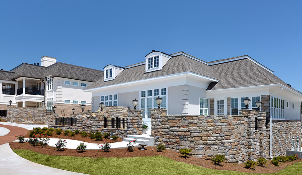Exterior view of a large, elegant retirement community building with stone walls, multiple windows, and a shingled roof under a clear blue sky. The landscaped area in front features green grass, mulch, and small bushes and flowers.