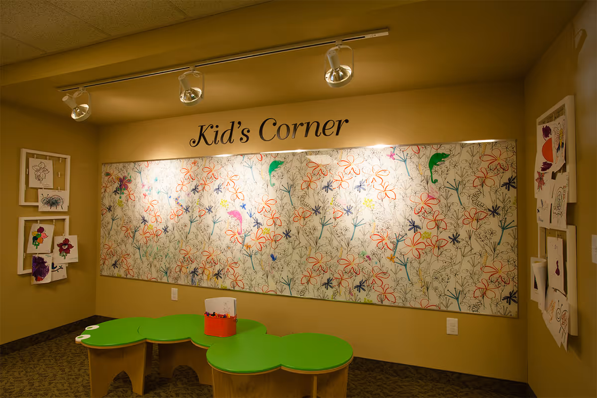 Indoor kids' activity corner with a 'Kid's Corner' sign, a large floral mural, children's artwork on the walls, and green child-sized tables.