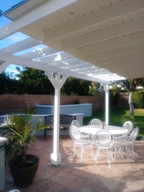 Outdoor patio area with a white pergola providing partial shade over a round white metal table and six matching chairs. There is a potted plant on the tiled floor and a brick wall with greenery and trees in the background.