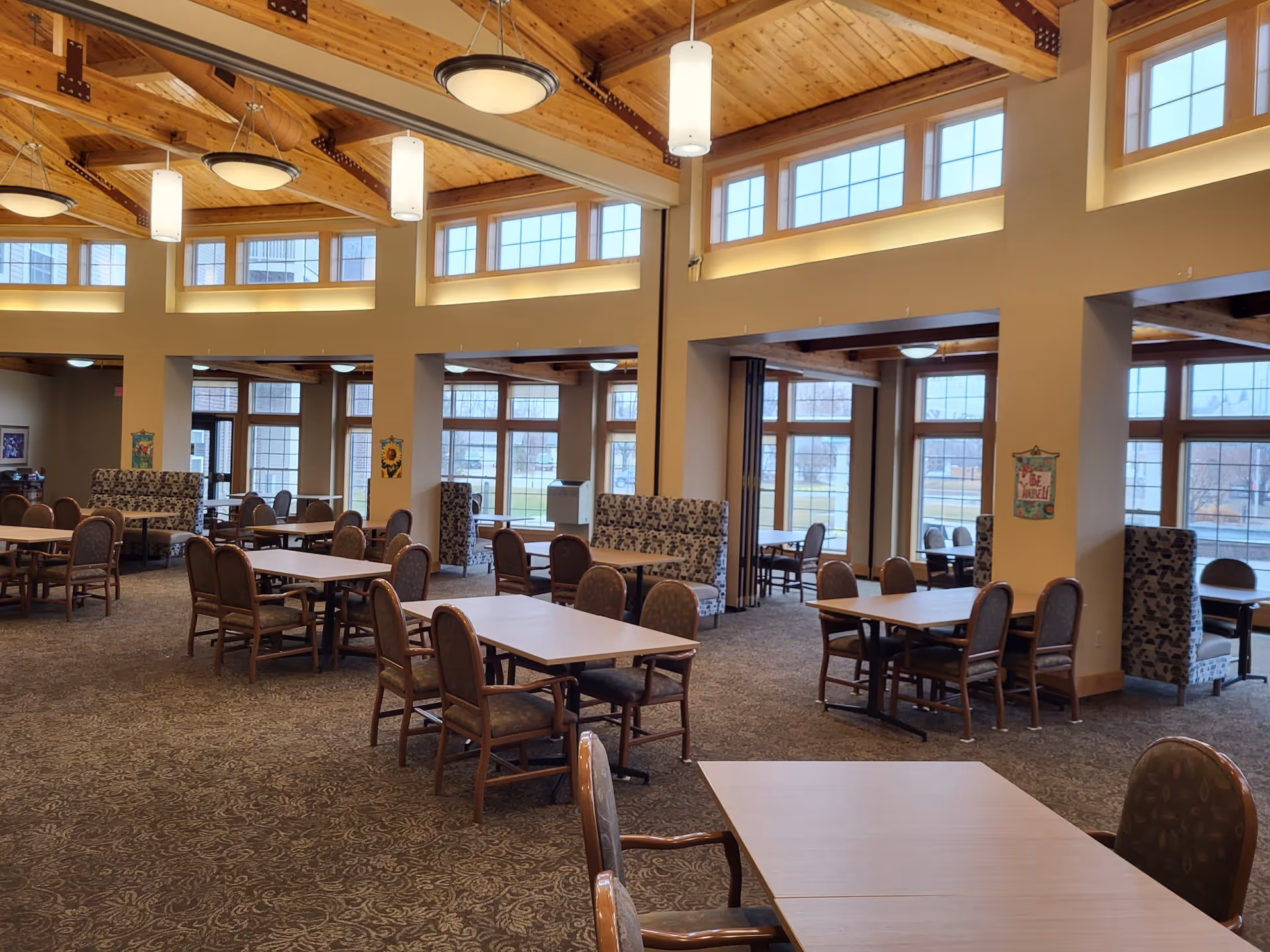 Spacious senior living dining area with multiple tables and chairs arranged neatly. The room features large windows allowing natural light to fill the space, high wooden ceilings with exposed beams, and pendant lighting fixtures. Upholstered booth seating is placed along the walls, and the carpet has a patterned design.