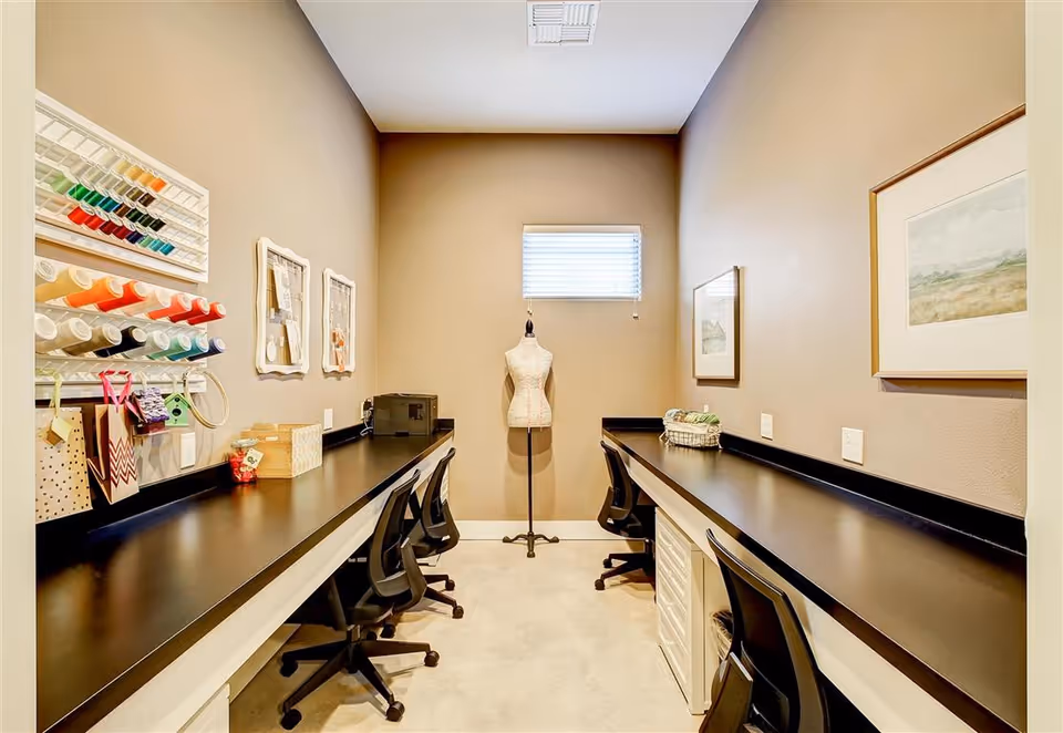 A narrow craft room with two long black countertops on either side, each with black office chairs. On the left wall, there are shelves holding colorful spools of thread and some hanging craft supplies. At the end of the room, there is a dress form mannequin in front of a small window with blinds. The walls are painted beige, and there are framed pictures on the right wall.
