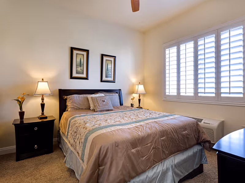 Bright bedroom with a made bed, two bedside tables with lamps, framed artwork above the headboard, and a window with shutters.