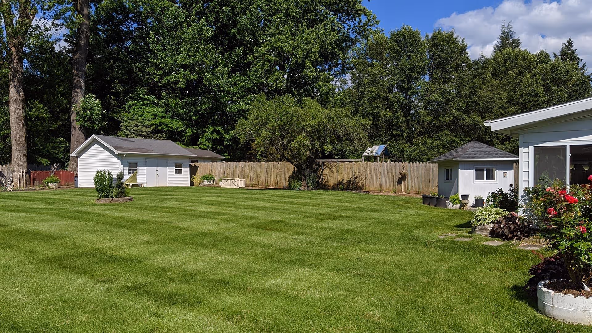 A large, well-maintained grassy backyard with a few small white buildings and a wooden fence in the background. There are trees behind the fence and some flower beds with blooming plants near the buildings. The sky is blue with some clouds.