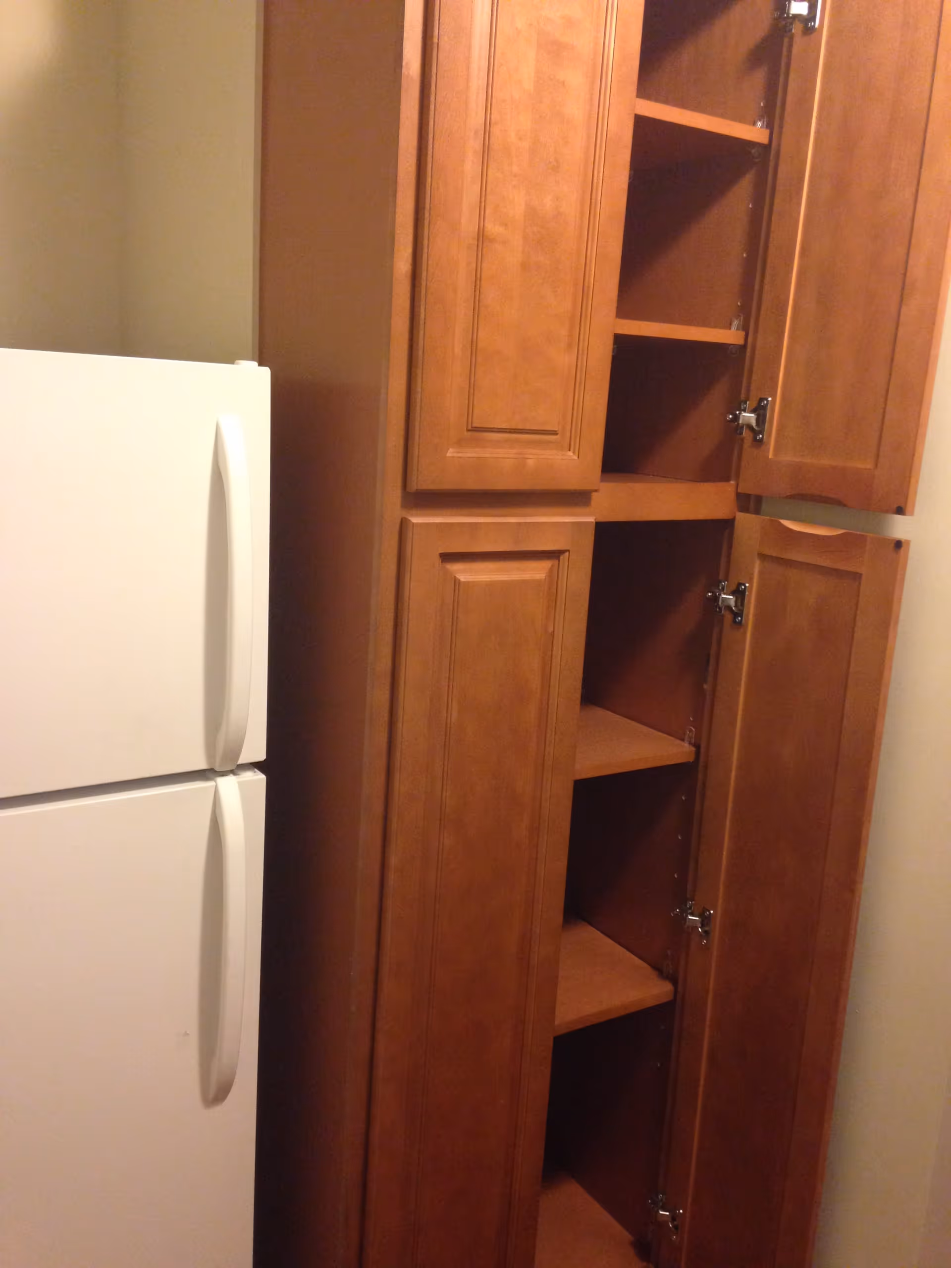 A white refrigerator next to a tall wooden cabinet with multiple shelves, some of the cabinet doors are open revealing empty shelves inside.