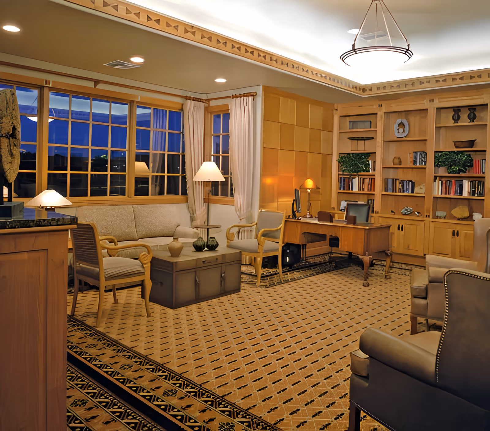 A cozy and well-lit living room with wooden furniture including a sofa, armchairs, a coffee table, a desk with a computer, and built-in bookshelves filled with books and decorative items. Large windows with curtains reveal a nighttime view outside. The room features a patterned carpet and warm lighting from lamps and ceiling lights.