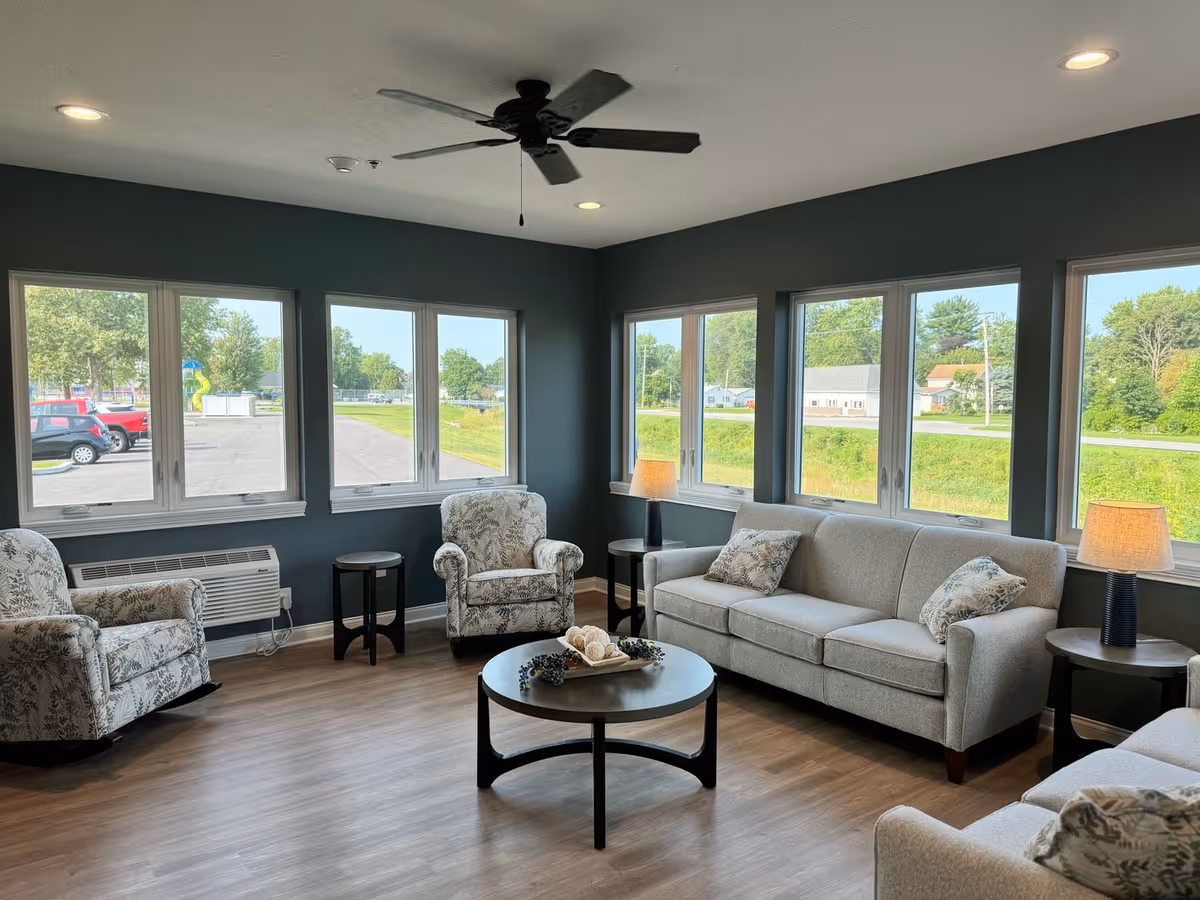 A bright and spacious living room with large windows showing an outdoor view. The room features a gray sofa with patterned pillows, two floral armchairs, a round coffee table with decorative items, two side tables with lamps, and a ceiling fan. The walls are painted dark gray and the floor has a wood finish.