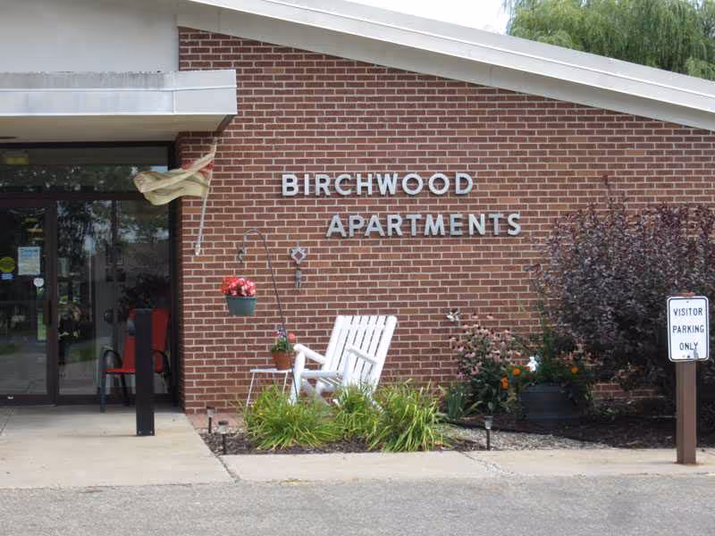 Brick building entrance labeled Birchwood Apartments with a white chair and garden in front.