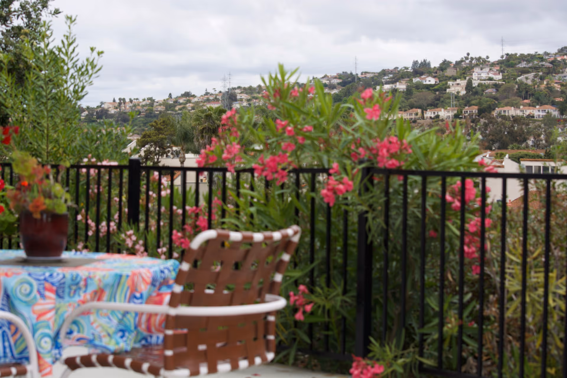Outdoor patio area with a table covered in a colorful tablecloth and a brown woven chair. The patio is surrounded by a black metal fence and lush greenery with pink flowers. In the background, there is a hillside with houses and trees under a cloudy sky.