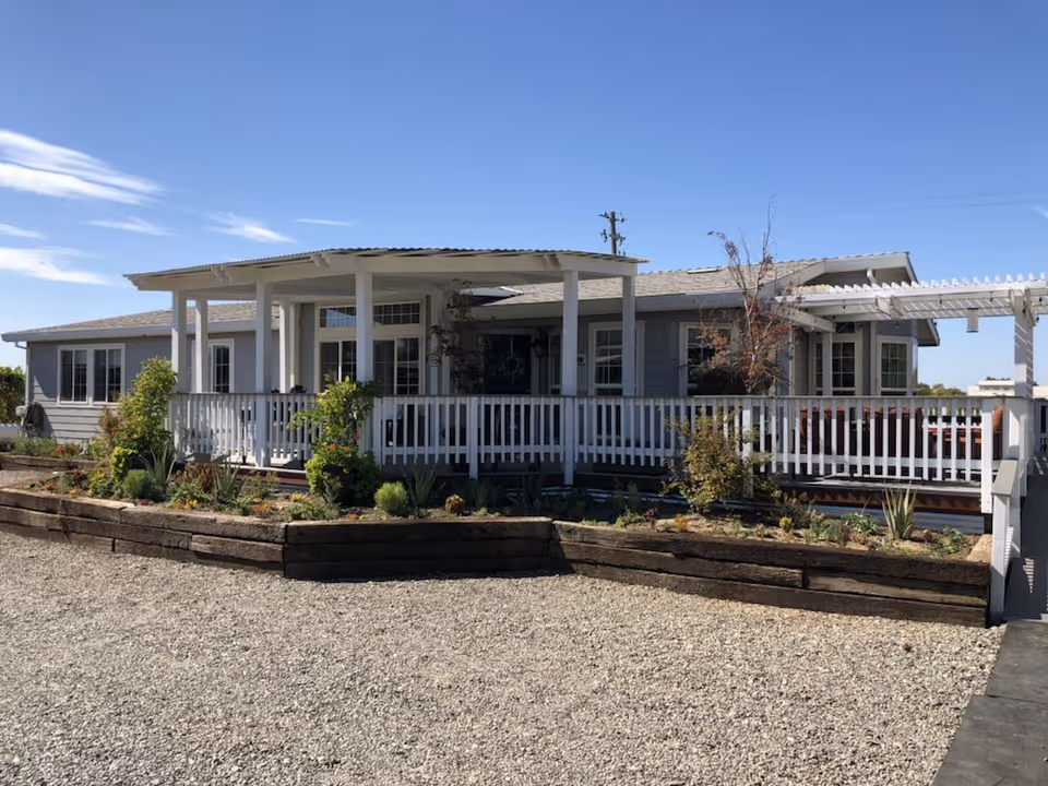 Single-story light-gray building with a covered porch, white railing and raised garden beds in front under a clear blue sky.