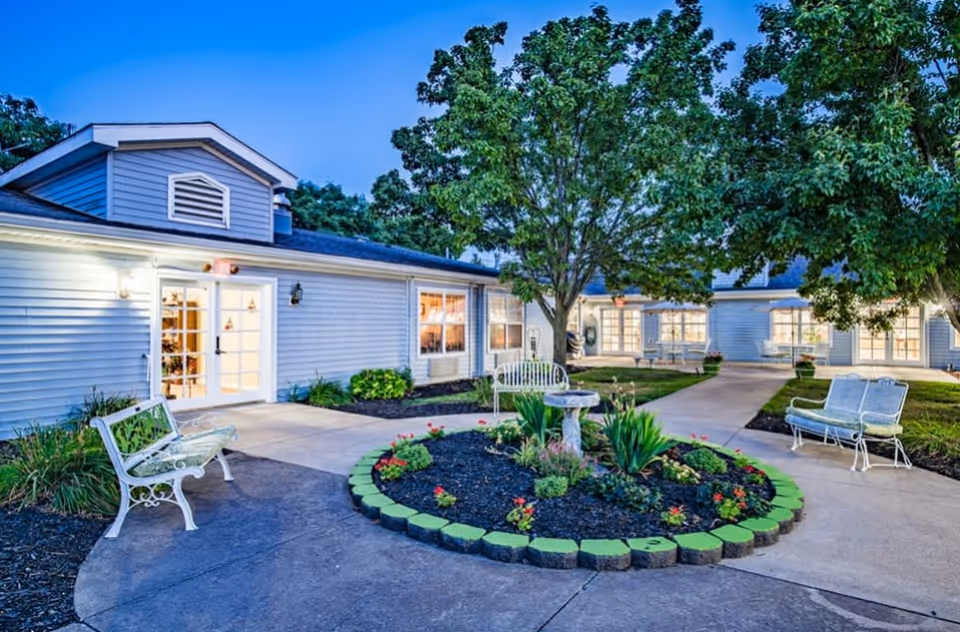 Outdoor courtyard area of Randall Residence of Tiffin featuring a circular flower bed with a birdbath in the center, surrounded by paved walkways, white benches, and trees. The building exterior is light-colored with multiple windows and glass doors, illuminated by outdoor lighting at dusk.