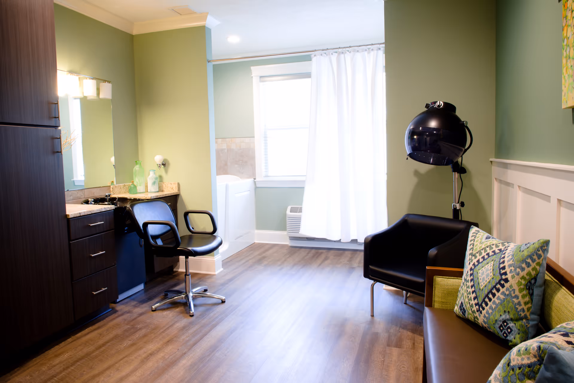 A well-lit room with light green walls and wood flooring featuring a black salon chair in front of a vanity with a mirror and countertop. To the right, there is a black hair dryer chair and a brown couch with colorful patterned pillows. A window with white curtains is in the background.