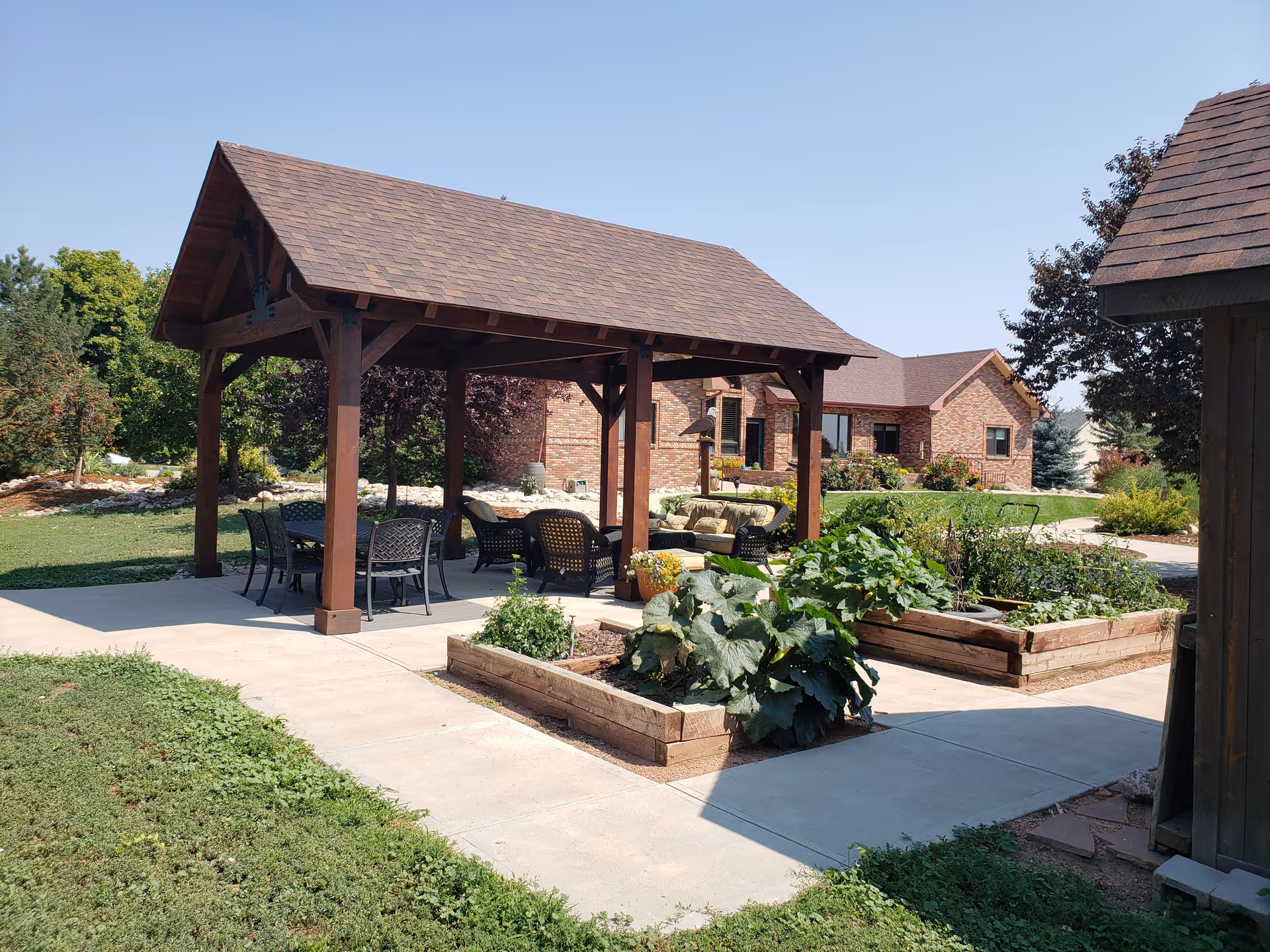 Outdoor patio area with a wooden pergola covering a seating arrangement including chairs and a table. Raised garden beds with green plants and vegetables are in the foreground. A brick building and trees are visible in the background under a clear blue sky.