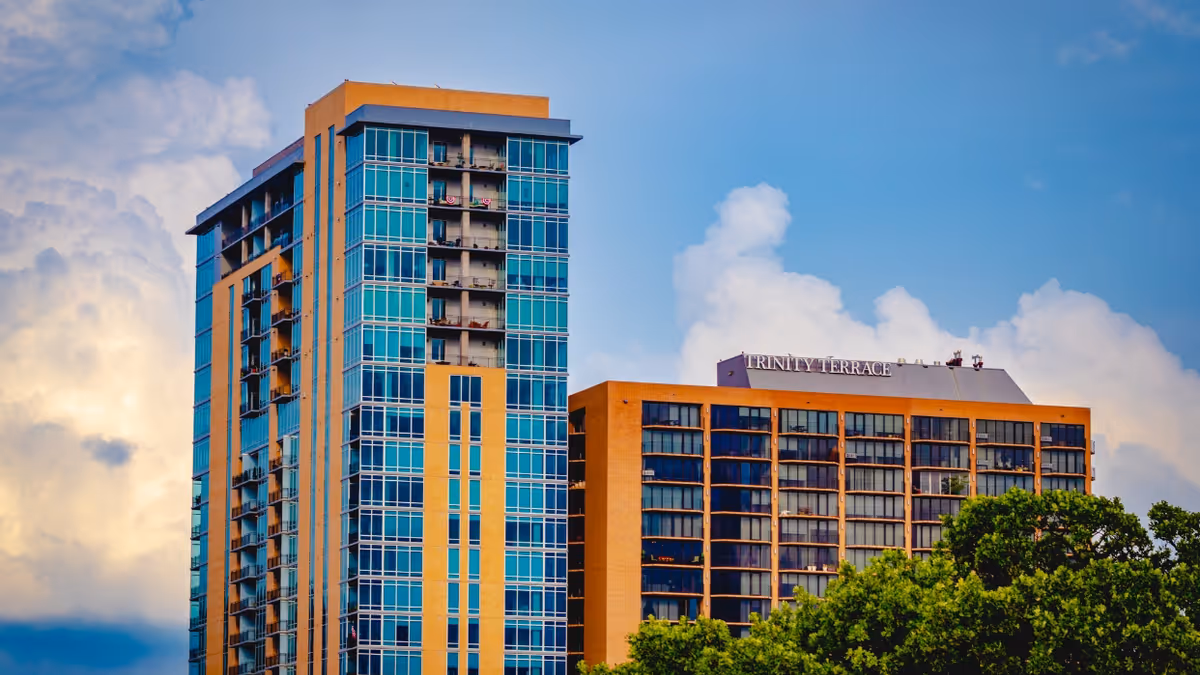 Exterior view of a modern multi-story senior living facility named Trinity Terrace, featuring two connected buildings with large windows and balconies, set against a partly cloudy blue sky with green trees in the foreground.