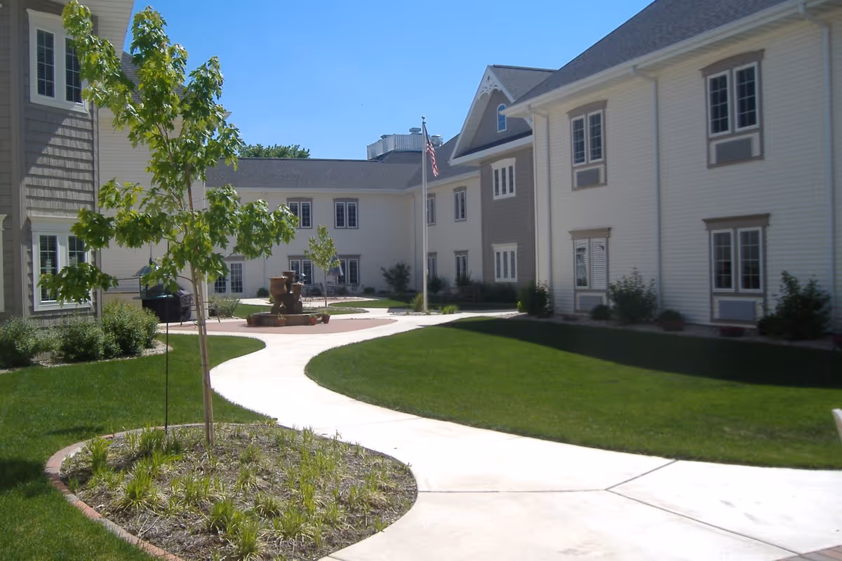 Sunlit courtyard with a winding concrete path, grassy lawns, a fountain, small trees, and surrounding two-story senior living buildings.