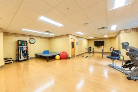 A spacious rehabilitation exercise room with light wood flooring and beige walls. The room contains exercise equipment including a treadmill, stationary bike, parallel bars for walking support, a therapy table, exercise balls, and a rack with various resistance bands and weights. A wall-mounted TV is visible in the background.