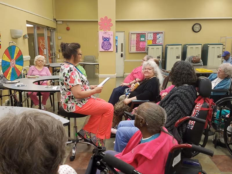 A group of elderly individuals seated in a room, some in wheelchairs, attentively listening to a woman in a floral top and pink pants who is reading from a paper. The room has yellow walls, a colorful spinning wheel, lockers, and bulletin boards in the background.