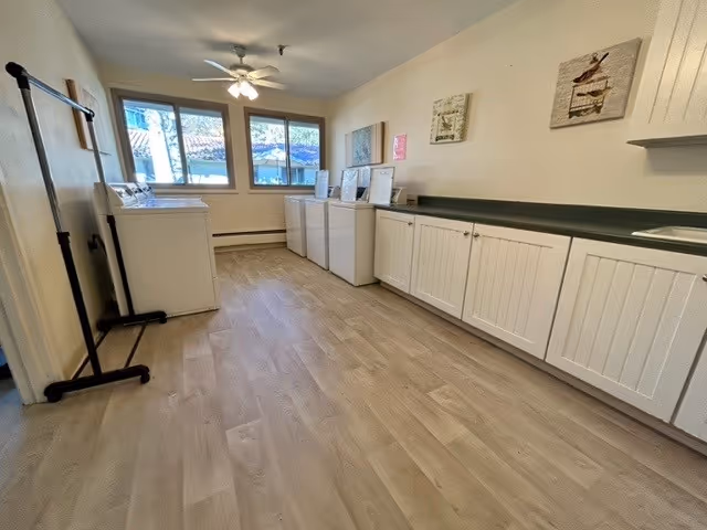 A bright laundry room with light wood flooring, white cabinets along the right wall, and three washing machines and dryers against the far wall under two windows. A ceiling fan with lights is mounted on the ceiling, and there are framed pictures hanging on the walls.