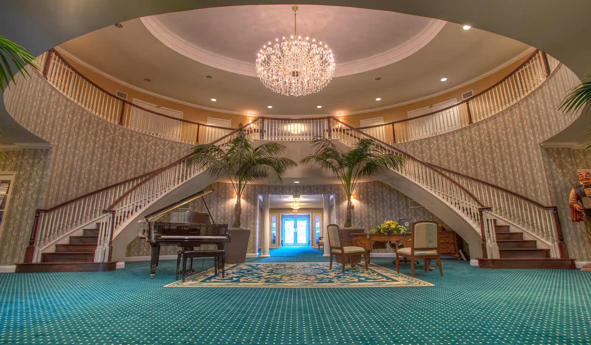 Spacious elegant lobby area with a grand double staircase curving upwards on both sides, a large crystal chandelier hanging from the ceiling, a black grand piano on the left, two tall potted palm plants, and a seating area with a wooden table and chairs on a patterned rug. The walls have a subtle floral wallpaper and the floor is covered with green carpet with small white dots.