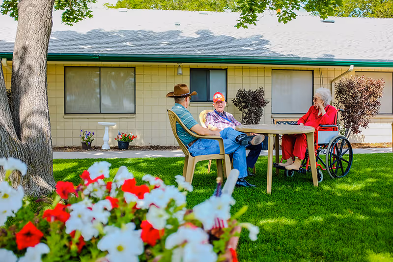 Three older adults—two seated in chairs and one in a wheelchair—talk around a table on a grassy courtyard outside a single-story nursing facility, with flowers in the foreground.