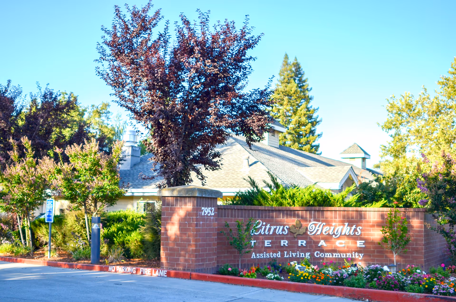 Brick entry sign reading "Citrus Heights Terrace Assisted Living Community" with flowers, trees and part of the building visible behind it.