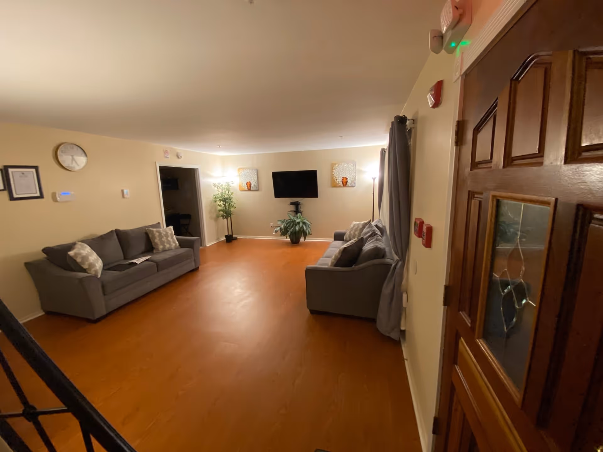 A cozy living room with two gray sofas facing each other, a wall-mounted flat screen TV, two decorative wall hangings, a potted plant on the floor, and a floor lamp. The room has wooden flooring, beige walls, and a wooden door with a glass panel on the right side.