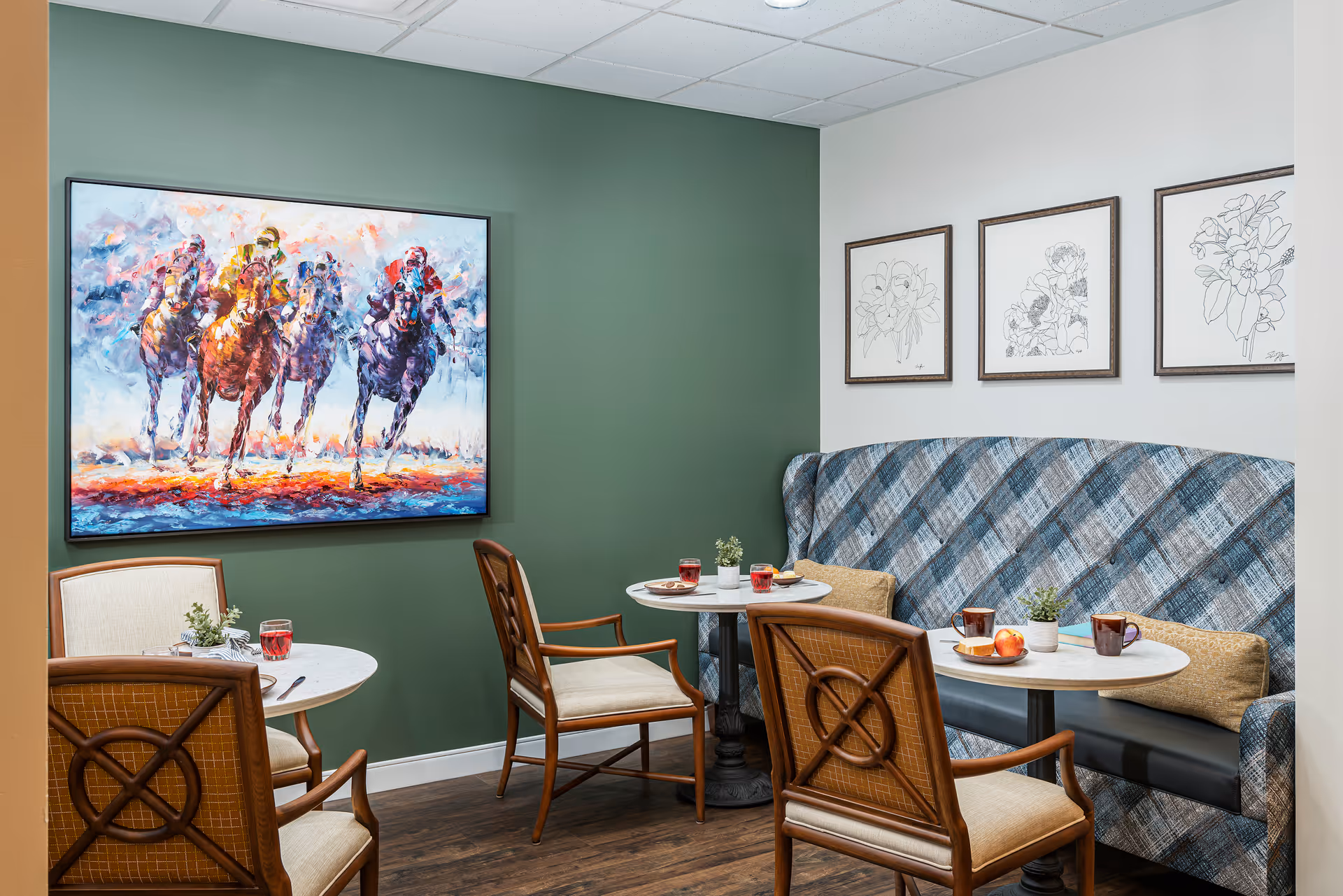 Small dining nook with round tables and chairs, a plaid upholstered banquette, colorful horse-racing painting and framed botanical sketches on the walls.