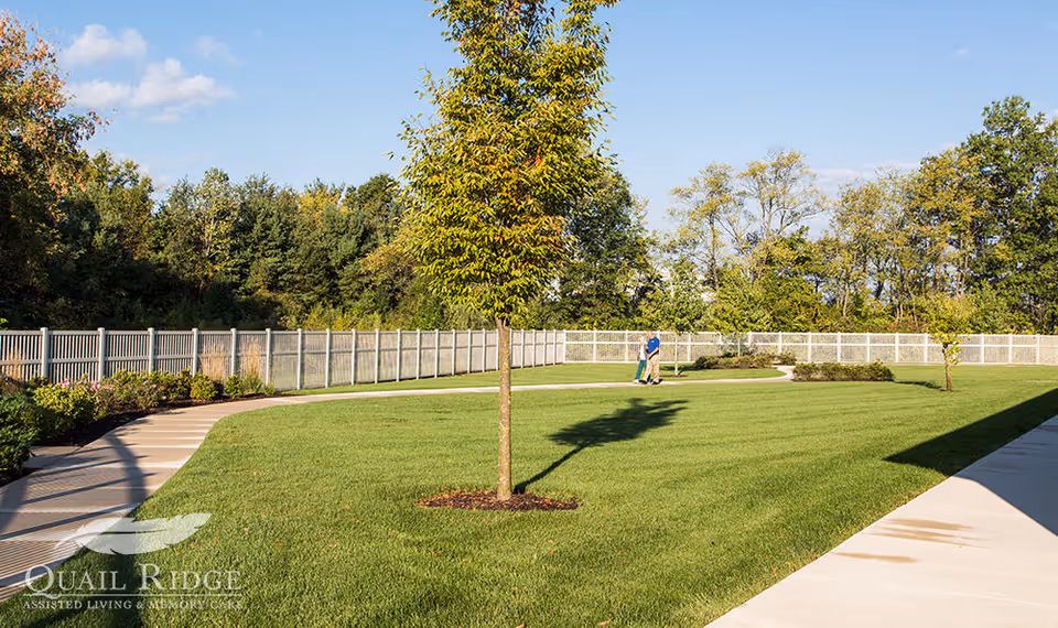 A spacious, well-maintained outdoor lawn area with a paved walking path curving around the grass. There are a few young trees planted in the lawn, and a white fence encloses the area. Two people are walking along the path in the background, and there are dense trees beyond the fence under a clear blue sky.