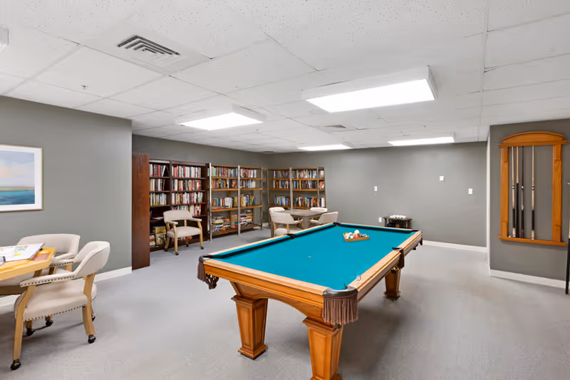 A recreational room with a pool table in the center, surrounded by chairs and small tables. There are bookshelves filled with books along the back wall and a rack holding pool cues mounted on the right wall. The room has gray walls, a carpeted floor, and bright overhead lighting.