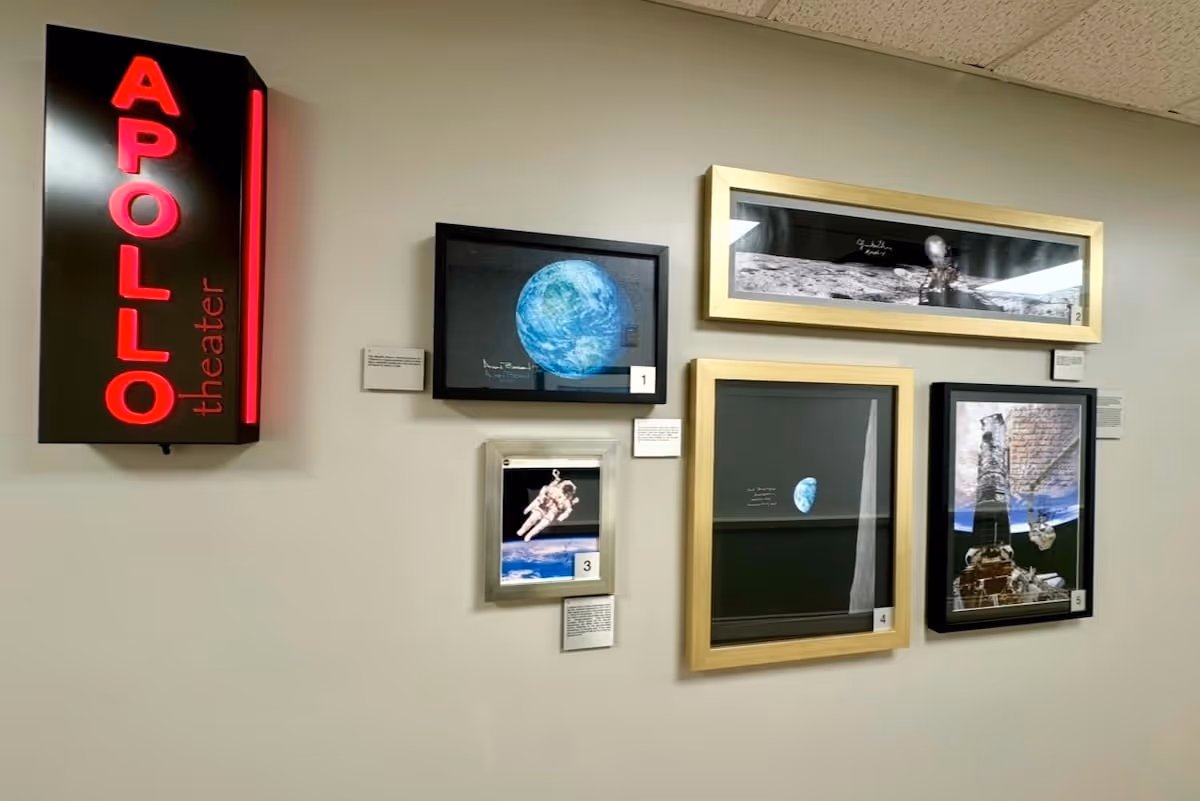 Wall display featuring a red illuminated sign that reads 'APOLLO theater' alongside framed photographs and artwork related to space exploration, including images of Earth, an astronaut, the moon's surface, and a space station.