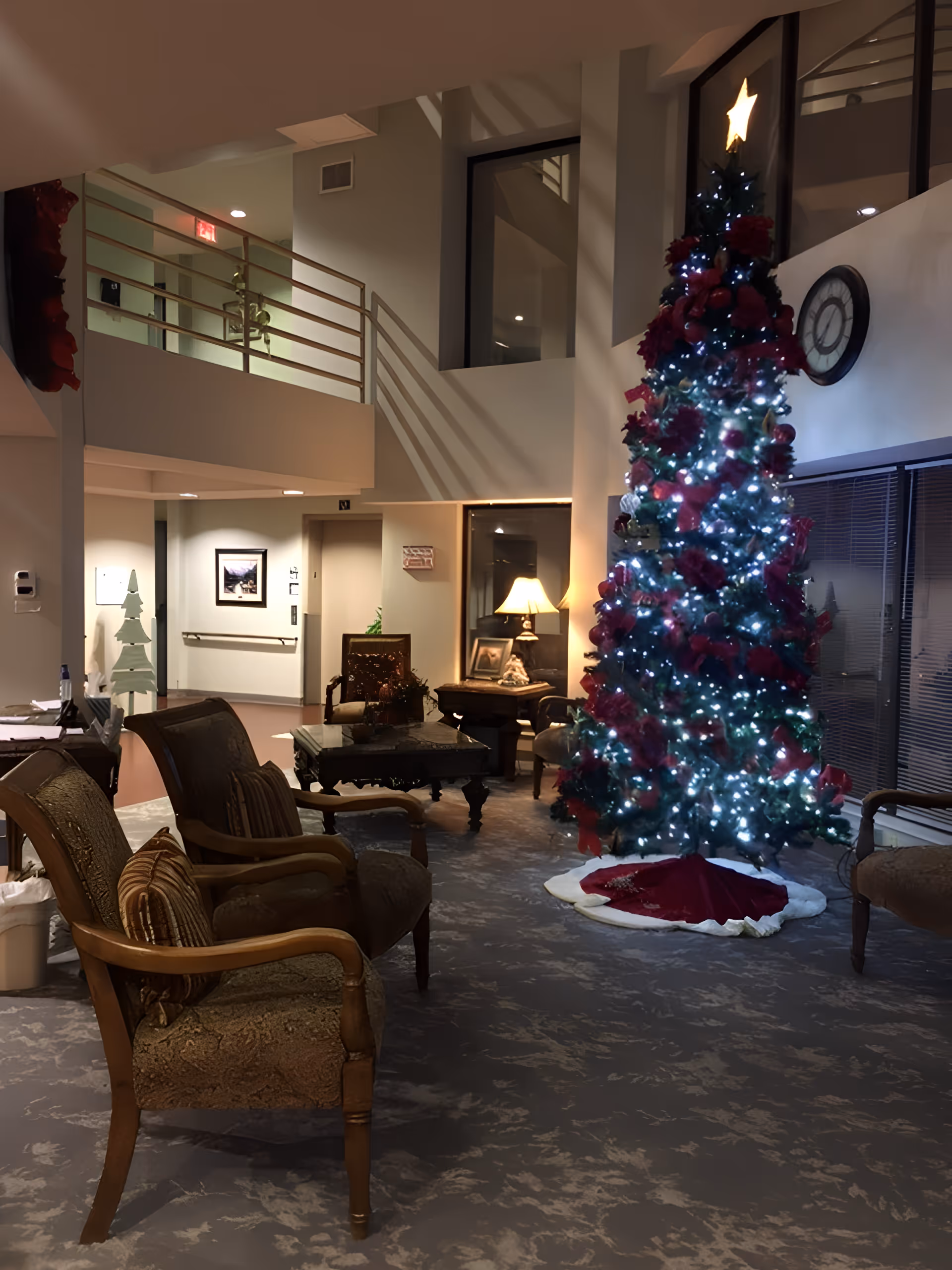A cozy indoor common area decorated for Christmas with a tall Christmas tree adorned with white lights, red flowers, and a star on top. The room features several upholstered chairs with wooden arms, a coffee table, a side table with a lamp, and a large wall clock. The space has high ceilings and a second-floor balcony with railings.