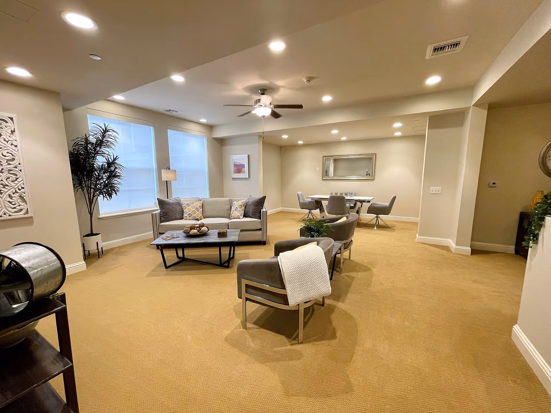 A spacious, well-lit living room area with beige carpet and neutral-colored walls. The room features a gray sofa with patterned cushions, two gray armchairs with a white throw blanket on one, a rectangular coffee table with decorative items, and a dining table with four chairs in the background. There are two large windows with blinds, a ceiling fan with lights, recessed ceiling lights, a tall potted plant, and framed artwork on the walls.