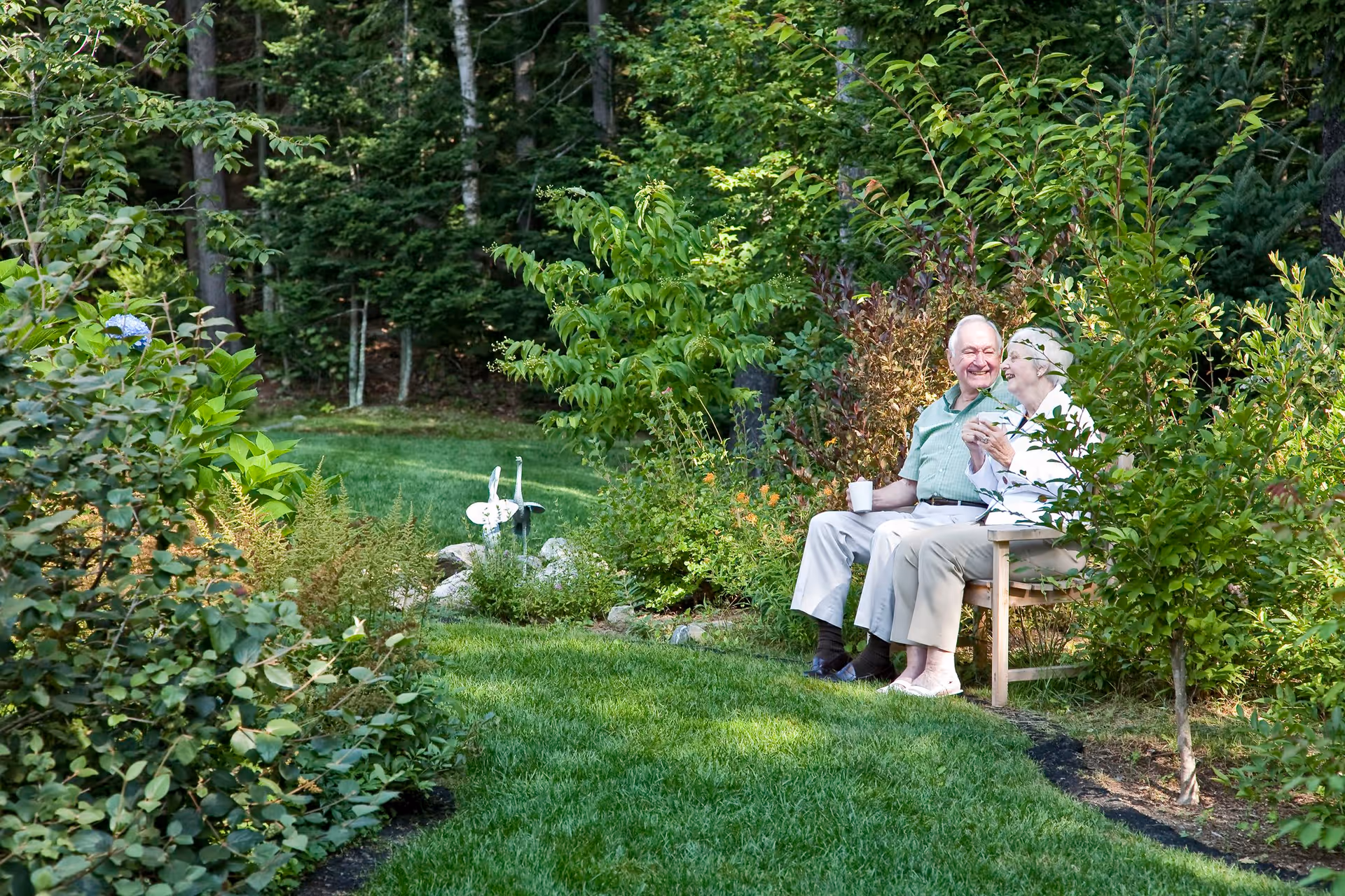 An elderly couple sitting on a wooden bench in a lush garden surrounded by green plants and trees, enjoying a peaceful moment together with cups in their hands.