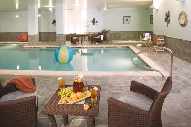 Indoor swimming pool area with a beach ball floating in the water. In the foreground, there is a small table with a pitcher of iced tea, two glasses with lemon slices, and a plate of fruit. Two wicker chairs with cushions are positioned around the table. The pool area has tiled flooring and walls, with some decorative wall art and a clock on the wall. There are also additional chairs and a small table in the background near the pool.
